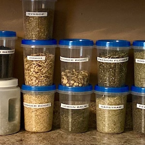 May include: A collection of clear plastic containers with blue lids, filled with various dried herbs. Labels identify each herb, including Calendula, Chamomile, Hyssop, Elder Berry, Hydrangea, Licorice Root, Passion Flower, Rosemary, Sarsaparilla, Ginger Root, Lemon Balm, Oatstraw, Peppermint, Sage, and Yellow Dock.