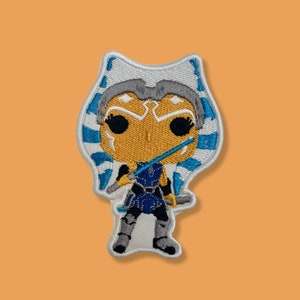 May include: An embroidered patch featuring a cartoon-style depiction of Ahsoka Tano, a Jedi Knight from the Star Wars universe. She is wearing her signature blue and white head-tails and wielding a blue lightsaber.