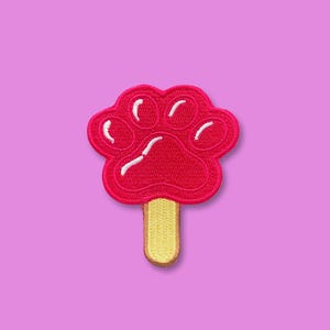 May include: Embroidered patch featuring a red paw print design on a yellow popsicle stick. The paw print has white accents and is set against a light purple background. This patch is suitable for pet lovers.