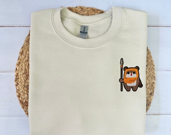 May include: A cream colored sweatshirt with a small embroidered image of a brown and orange character holding a spear. The character is wearing a brown hooded robe and has a yellow face with orange eyes. The text "YUTOPIA CREATIONS" is printed on the bottom of the sweatshirt.