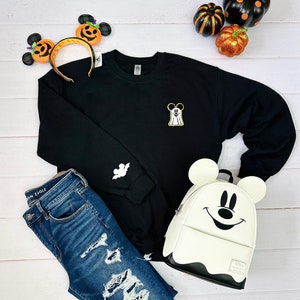 May include: Black sweatshirt with a white ghost Mickey Mouse design on the front and a white ghost Mickey Mouse design on the sleeve.  The sweatshirt is paired with blue jeans, a white ghost Mickey Mouse backpack, and a headband with Mickey Mouse ears and a pumpkin design.