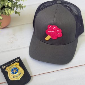 May include: A charcoal gray trucker hat with a pink popsicle patch on the front. The hat has a black mesh back and a curved brim. A black leather police officer badge is next to the hat.