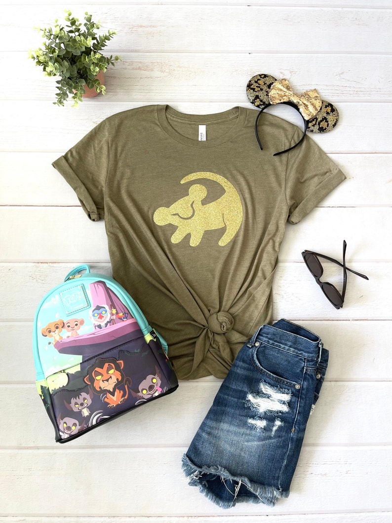 Puede incluir: Una camiseta verde con una silueta de le&oacute;n dorada con purpurina, una mochila azul y verde con estampado de el rey le&oacute;n, pantalones cortos de mezclilla y una diadema negra con orejas doradas.