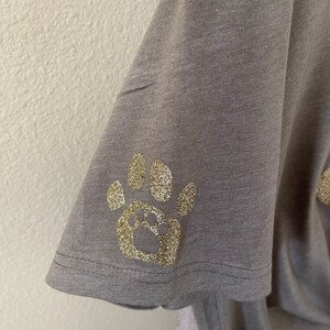 Puede incluir: Una camiseta gris con un dise&ntilde;o de huella de pata de purpurina dorada.
