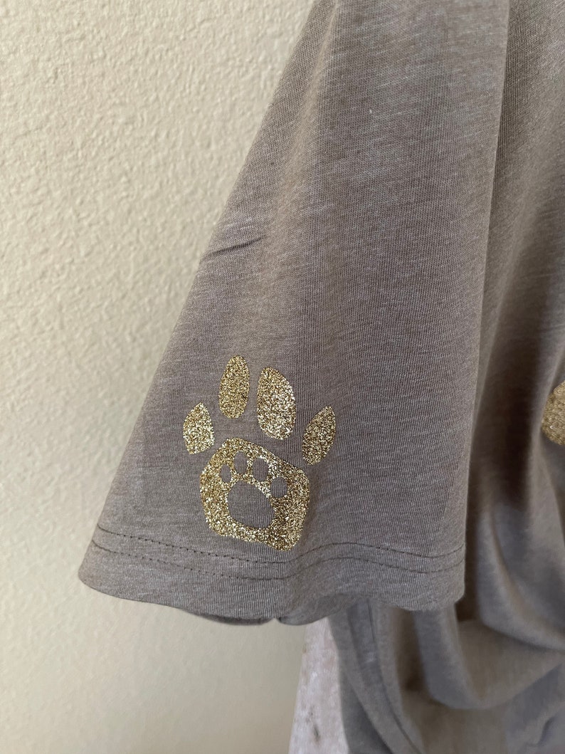 Puede incluir: Una camiseta gris con un dise&ntilde;o de huella de pata de purpurina dorada.