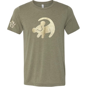 Puede incluir: Una camiseta verde con un contorno dorado de purpurina de Simba de El Rey Le&oacute;n. La camiseta tiene una huella de pata en la manga izquierda.