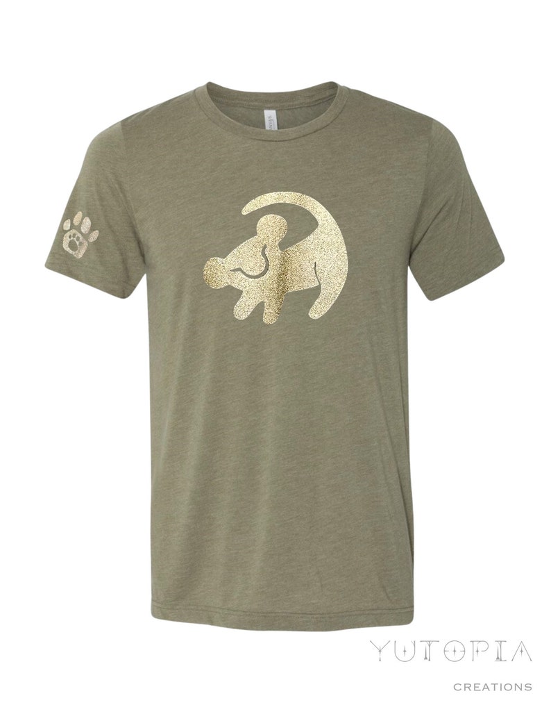 Puede incluir: Una camiseta verde con un contorno dorado de purpurina de Simba de El Rey Le&oacute;n. La camiseta tiene una huella de pata en la manga izquierda.