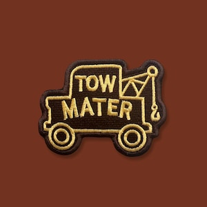 May include: Embroidered patch featuring a cartoon tow truck design. The patch is black with gold-colored stitching outlining the truck and the words "TOW MATER". The patch has a brown background.