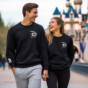 Pu&ograve; includere: Felpe nere con logo Disney bianco e scritta "Est. 1971". Una coppia sorridente indossa queste felpe. Sullo sfondo si vede un castello.