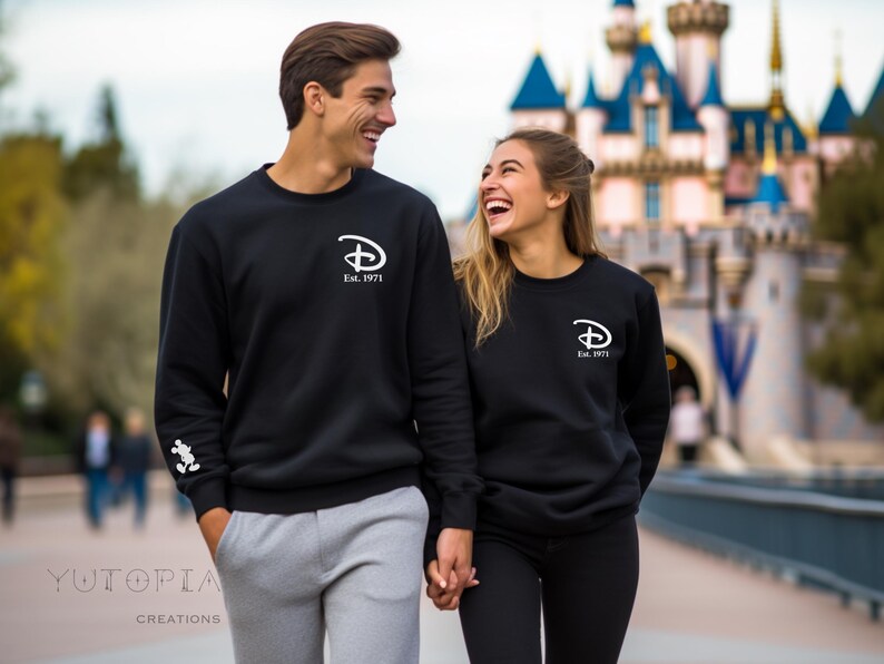 Pu&ograve; includere: Felpe nere con logo Disney bianco e scritta "Est. 1971". Una coppia sorridente indossa queste felpe. Sullo sfondo si vede un castello.