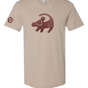 Puede incluir: Una camiseta beige con una silueta de le&oacute;n rey de purpurina marr&oacute;n en la parte delantera. La manga izquierda tiene una huella de pata de purpurina marr&oacute;n.
