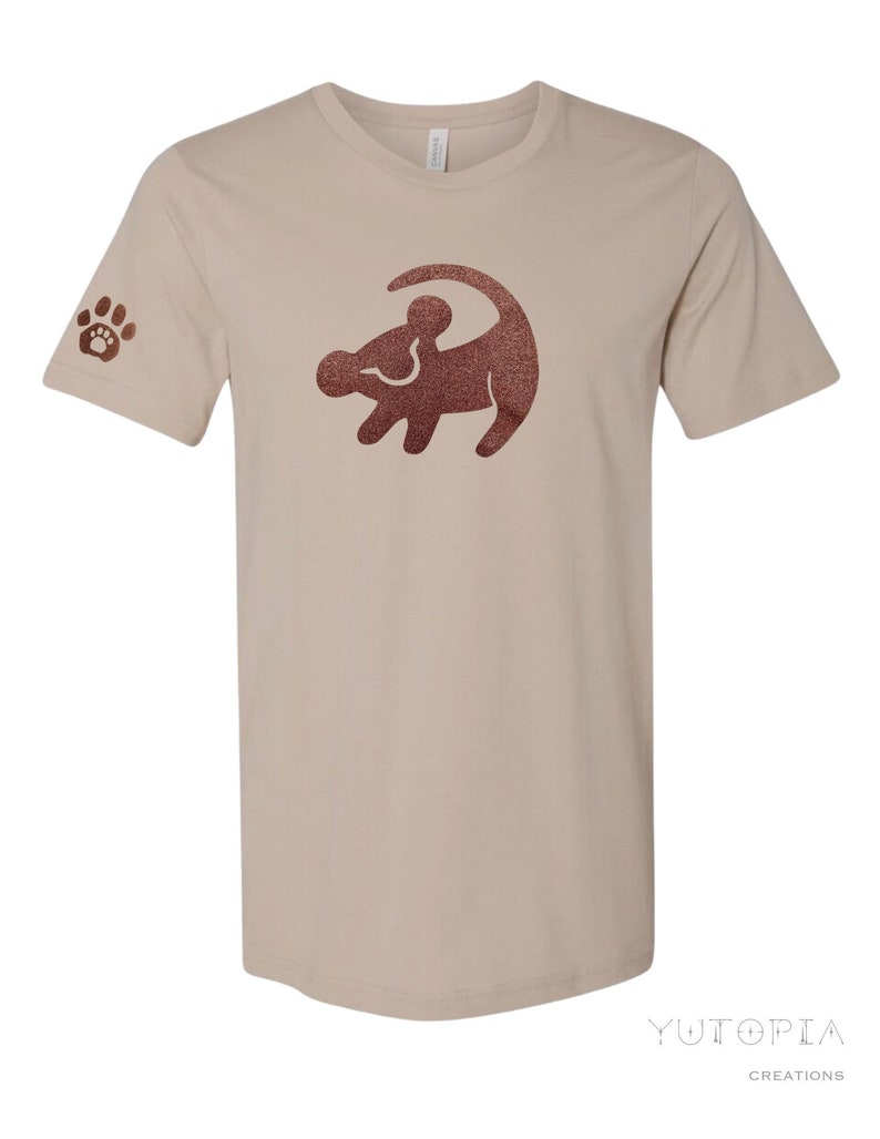 Puede incluir: Una camiseta beige con una silueta de le&oacute;n rey de purpurina marr&oacute;n en la parte delantera. La manga izquierda tiene una huella de pata de purpurina marr&oacute;n.