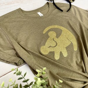 Puede incluir: Una camiseta verde con un dise&ntilde;o de huella de pata de le&oacute;n de purpurina dorada. La camiseta est&aacute; hecha de un material suave y c&oacute;modo.