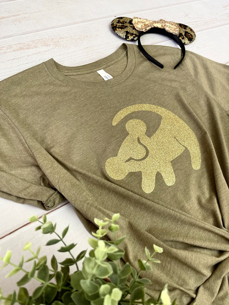 Puede incluir: Una camiseta verde con un dise&ntilde;o de huella de pata de le&oacute;n de purpurina dorada. La camiseta est&aacute; hecha de un material suave y c&oacute;modo.