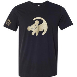 Puede incluir: Camiseta negra con un contorno de purpurina dorada de Simba de El Rey Le&oacute;n. La camiseta tiene una huella de pata en la manga.