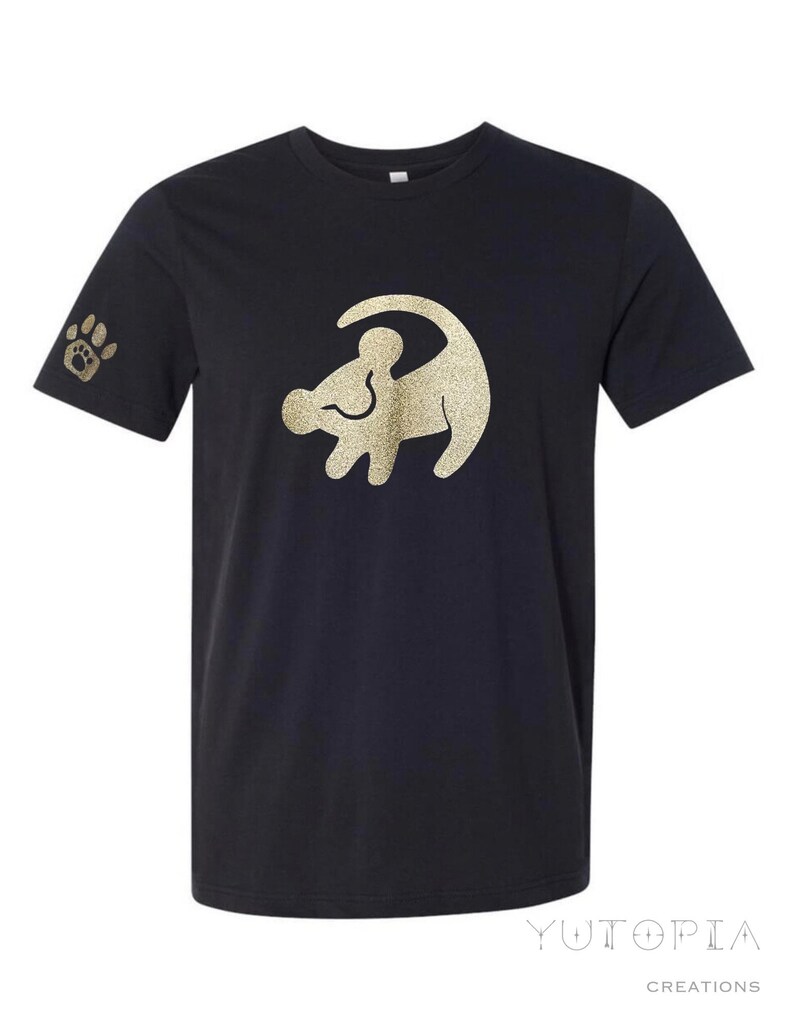 Puede incluir: Camiseta negra con un contorno de purpurina dorada de Simba de El Rey Le&oacute;n. La camiseta tiene una huella de pata en la manga.