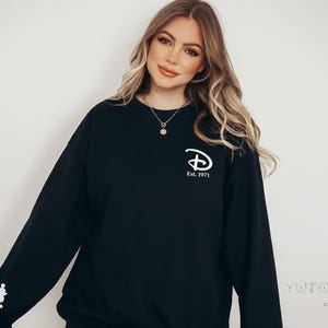 Pu&ograve; includere: Felpa nera con il logo Disney e la scritta "Est. 1971" in bianco. Una grafica di Mickey Mouse bianca &egrave; sulla manica sinistra. La felpa &egrave; in stile casual.