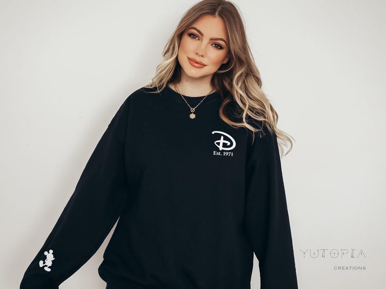 Pu&ograve; includere: Felpa nera con il logo Disney e la scritta "Est. 1971" in bianco. Una grafica di Mickey Mouse bianca &egrave; sulla manica sinistra. La felpa &egrave; in stile casual.