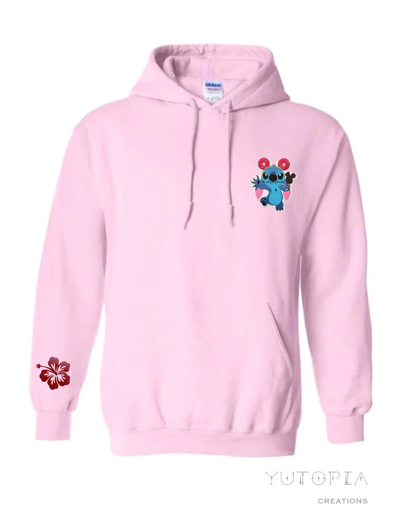 May include: Light pink hoodie with a front pocket and drawstring. Features a blue cartoon character with donuts and a hibiscus flower on the sleeve. The brand name "Yutopia Creations" is printed at the bottom.