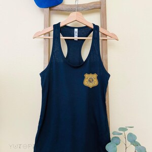 K&ouml;nnte beinhalten: Ein marineblaues Tanktop mit einem goldfarbenen Polizeiausweis auf der Brust. Dar&uuml;ber eine blaue Baseballkappe mit einer Cartoonfigur und einem Polizeiausweis. Das Tanktop h&auml;ngt an einem Holzb&uuml;gel.