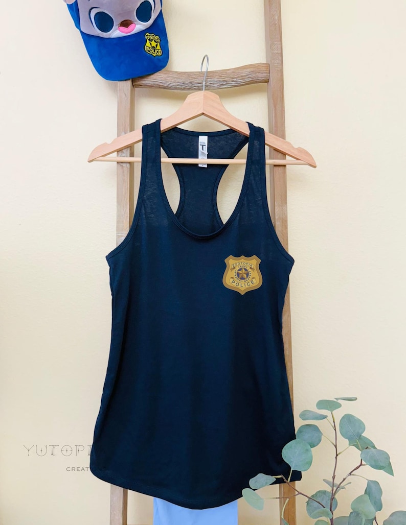 K&ouml;nnte beinhalten: Ein marineblaues Tanktop mit einem goldfarbenen Polizeiausweis auf der Brust. Dar&uuml;ber eine blaue Baseballkappe mit einer Cartoonfigur und einem Polizeiausweis. Das Tanktop h&auml;ngt an einem Holzb&uuml;gel.