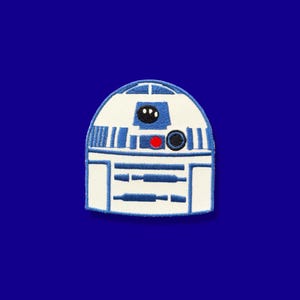 May include: Embroidered patch featuring the iconic R2-D2 droid from Star Wars. The patch has a white background with blue and black details, including a red circular accent. The design is set against a solid blue background.