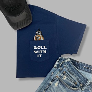 May include: A navy blue t-shirt with a pocket featuring a white and orange BB-8 character and the text "ROLL WITH IT" in white.
