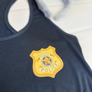K&ouml;nnte beinhalten: Ein marineblaues Tanktop mit einem gold-wei&szlig;en Polizeiausweis mit der Aufschrift "Zootopia Police". Im Hintergrund befindet sich ein graues Hasenohren-Stirnband. Das Tanktop ist aus einem weichen Material gefertigt.