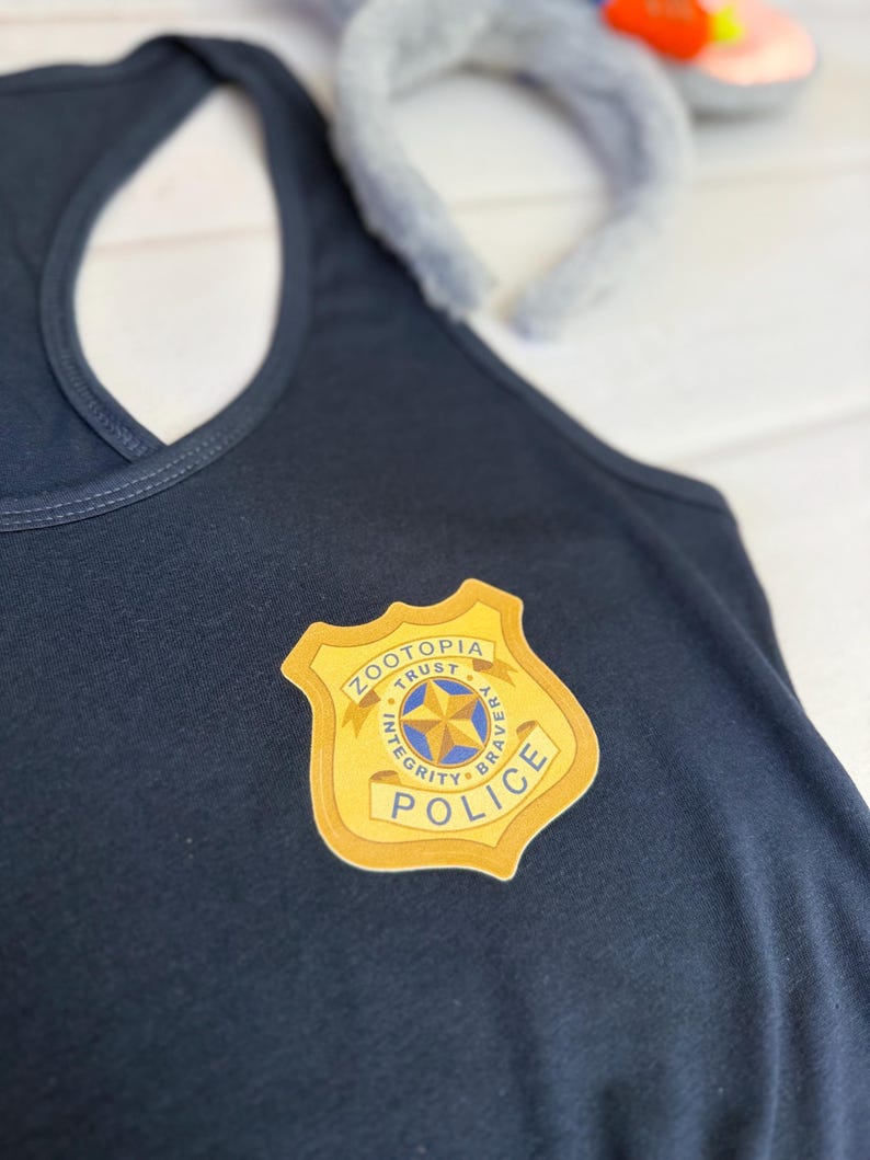 K&ouml;nnte beinhalten: Ein marineblaues Tanktop mit einem gold-wei&szlig;en Polizeiausweis mit der Aufschrift "Zootopia Police". Im Hintergrund befindet sich ein graues Hasenohren-Stirnband. Das Tanktop ist aus einem weichen Material gefertigt.