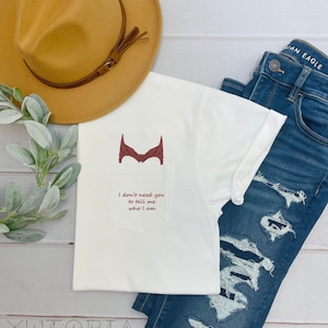 May include: White t-shirt with a red superhero symbol on the pocket and the text "I don't need you to tell me who I am".