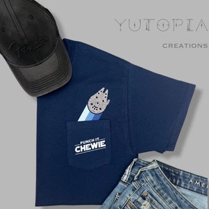 May include: A navy blue t-shirt with a pocket featuring a Millennium Falcon graphic and the text "Punch It, Chewie". The t-shirt is paired with blue jeans and a black baseball cap with the word "Fender" embroidered on it.
