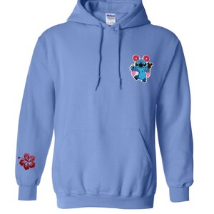 May include: A light blue hooded sweatshirt with a front pocket and drawstring. The left sleeve features a red hibiscus flower. The chest has an embroidered cartoon character holding a donut and ice cream. The brand name "Yutopia Creations" is printed at the bottom.