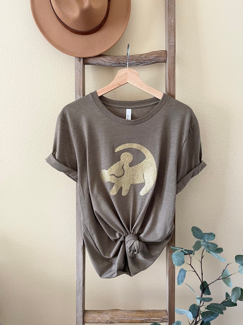 Puede incluir: Una camiseta marr&oacute;n con un contorno de purpurina dorada de Simba de El Rey Le&oacute;n.