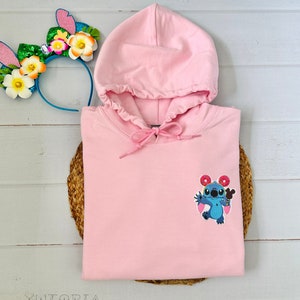 May include: A light pink hooded sweatshirt with a drawstring, featuring an embroidered cartoon character with donuts. A colorful floral and bunny ear headband rests nearby. The sweatshirt is folded on a woven mat.