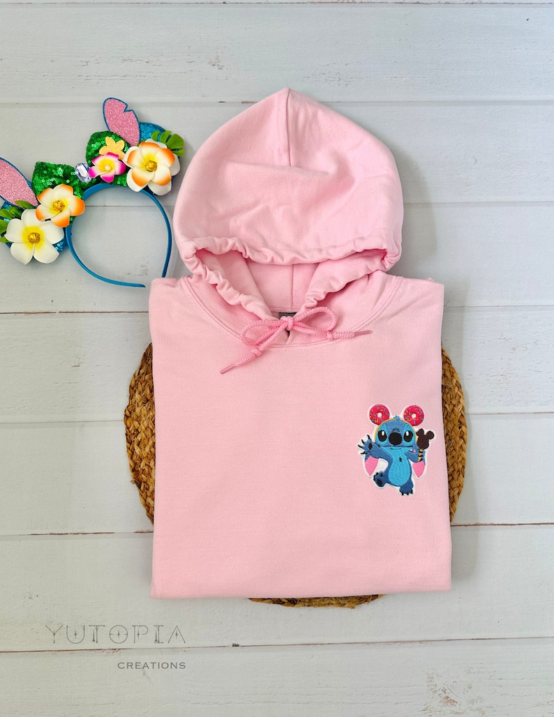 May include: A light pink hooded sweatshirt with a drawstring, featuring an embroidered cartoon character with donuts. A colorful floral and bunny ear headband rests nearby. The sweatshirt is folded on a woven mat.