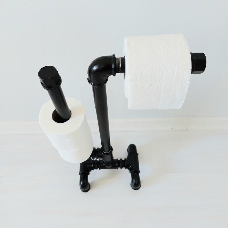 以下が含まれることがあります： 黒い金属製のトイレットペーパーホルダーに、トイレットペーパーが2ロール巻かれている。ホルダーは工業用パイプ継手でできており、素朴な外観です。