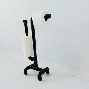 以下が含まれることがあります： 黒い金属製のトイレットペーパーホルダーに、トイレットペーパーが2ロール巻かれている。ホルダーは工業用パイプ継手で作られており、モダンなデザインである。