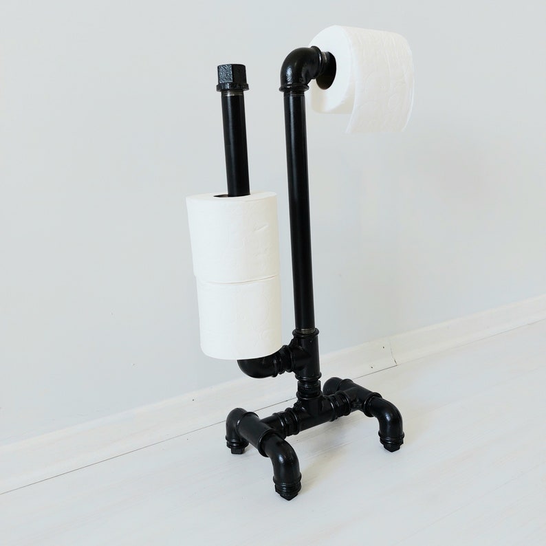 以下が含まれることがあります： 黒い金属製のトイレットペーパーホルダーに、トイレットペーパーが2ロール巻かれています。ホルダーは工業用パイプ継手で作られており、素朴な外観をしています。