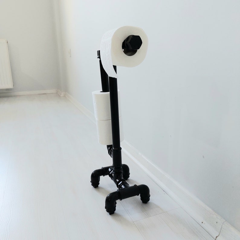 以下が含まれることがあります： 黒い金属製のトイレットペーパーホルダーに、トイレットペーパーが2ロール巻かれている。ホルダーは4本の脚で支えられている