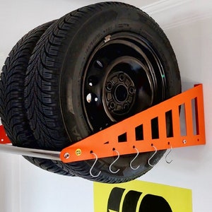 Könnte beinhalten: Ein orangefarbener Reifenhalter aus Metall, der an einer weißen Wand montiert ist, mit zwei schwarzen Reifen, die daran hängen. Der Halter hat eine silberne Stange und Haken.
