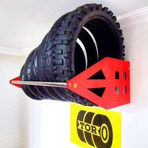 May include: Red metal tire rack mounted on a white wall with four black tires stacked on it. The rack has a silver metal bar and a logo that says "TORO" on a yellow sticker below the rack.