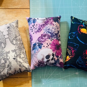 May include: Three decorative pillows with different patterns. The first pillow has a black and white floral design. The second pillow has a pink floral design with a skull in the center. The third pillow has a black background with a rainbow skull design.