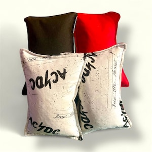 May include: Four decorative throw pillows. Two pillows are white with black text that says "AC/DC" and black speckles. One pillow is black and one pillow is red.