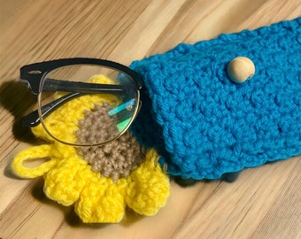 May include: A blue crocheted glasses case with a wooden button closure. The case is decorated with a yellow crocheted sunflower.