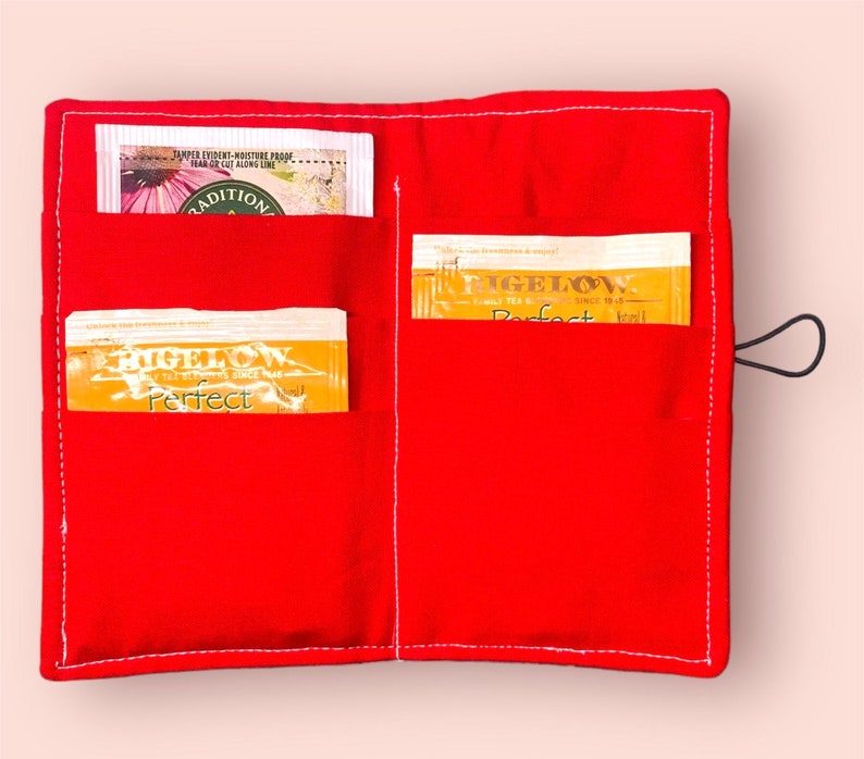 Puede incluir: Porta-bolsitas de t&eacute; de tela roja con dos bolsillos. Los bolsillos contienen bolsitas de t&eacute; con el texto "Figelow Perfect" en ellos.