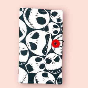 Puede incluir: Cartera de tela negra y blanca con un patr&oacute;n repetido de la cara de Jack Skellington de la pel&iacute;cula Pesadilla antes de Navidad. La cartera tiene un cierre de bot&oacute;n rojo.