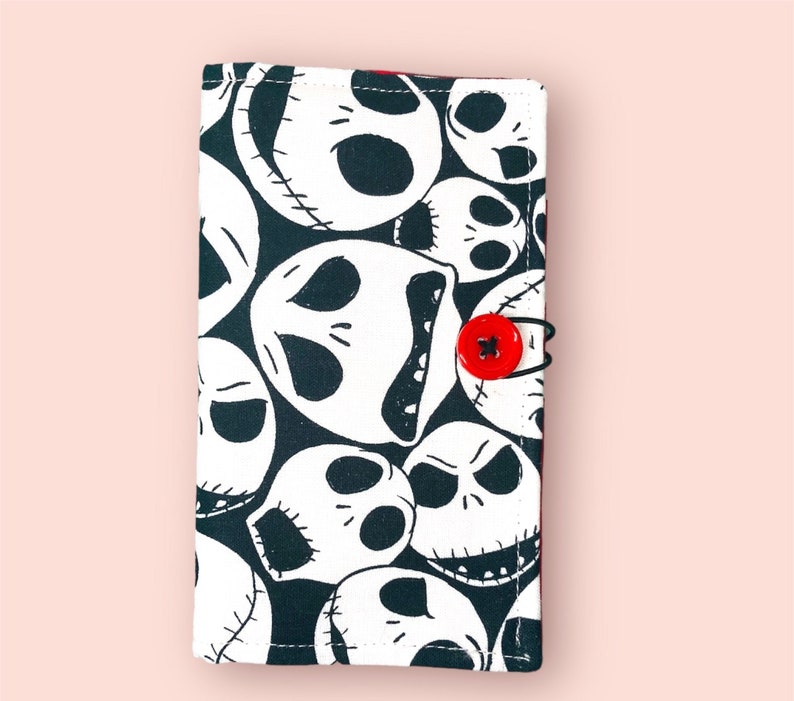 Puede incluir: Cartera de tela negra y blanca con un patr&oacute;n repetido de la cara de Jack Skellington de la pel&iacute;cula Pesadilla antes de Navidad. La cartera tiene un cierre de bot&oacute;n rojo.