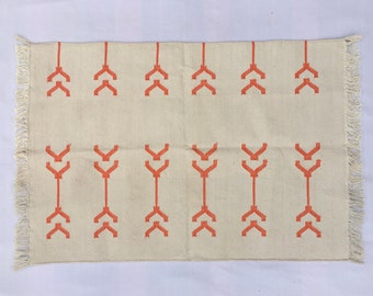 May include: Cream-colored rectangular rug with a repeating pattern of orange arrow-like shapes. The rug has a fringed edge on the short sides. The design is symmetrical and geometric, suitable for home decor.