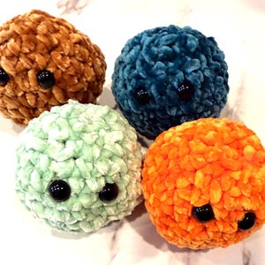 May include: Four plush, round toys in shades of brown, teal, mint green, and orange. Each has two large, black, button eyes. The toys are made of a soft, textured material. They are arranged on a light-coloured surface.