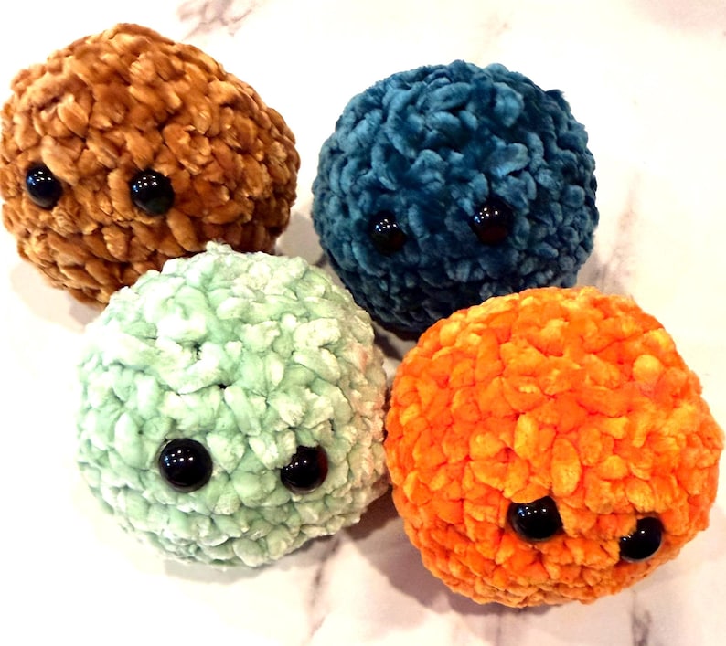 May include: Four plush, round toys in shades of brown, teal, mint green, and orange. Each has two large, black, button eyes. The toys are made of a soft, textured material. They are arranged on a light-coloured surface.